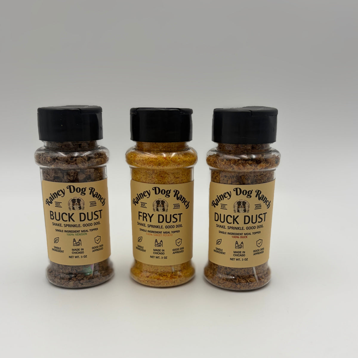 Three clear plastic bottles of Fancy Dog Ranch dog seasoning stands on a white studio background. The bottles contain brown and golden-brown spice dusts labeled Buck Dust, Fry Dust, and Duck Dust, with black caps.