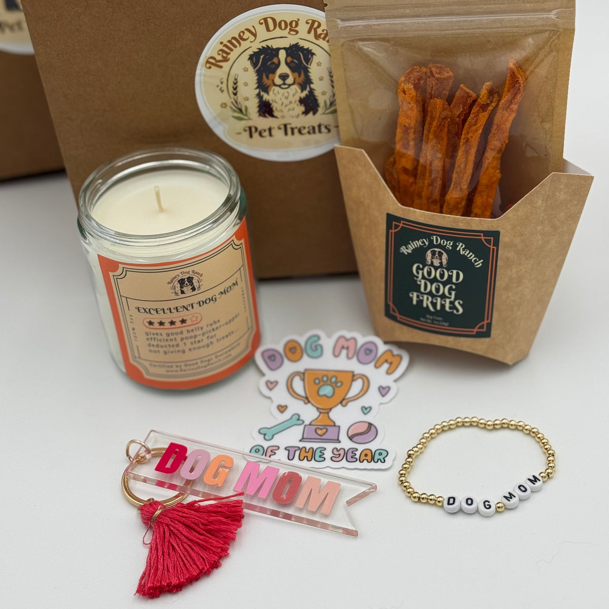 A curated gift set featuring a jar candle, a bag of dog treats, and several pet-themed accessories including a tassel keychain, a beaded bracelet, and stickers, arranged on a white surface with a brown paper bag in the background.