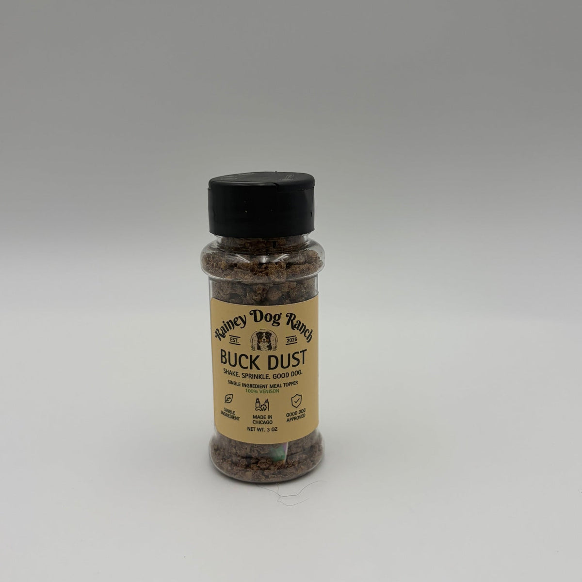 A small clear plastic bottle of Rainey Dog Ranch Buck Dust dog food meal topper with a black cap and a beige label, shown against a plain light gray background.