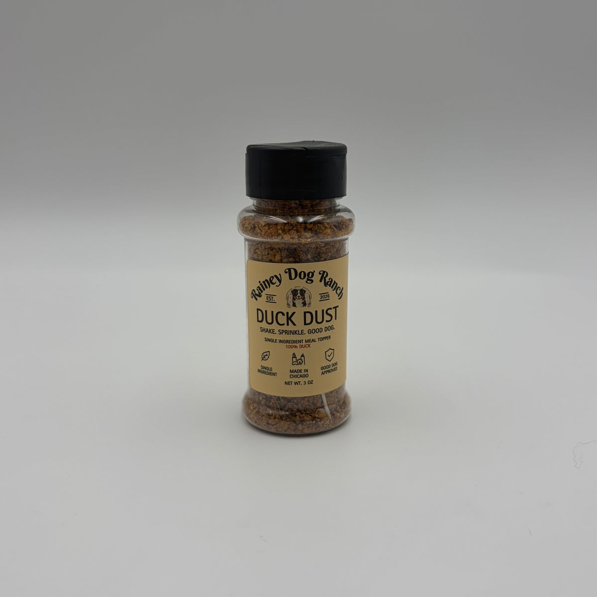 A small clear plastic bottle of Rainey Dog Ranch Duck Dust dog food meal topper with a black cap and tan label, set against a neutral gray background.