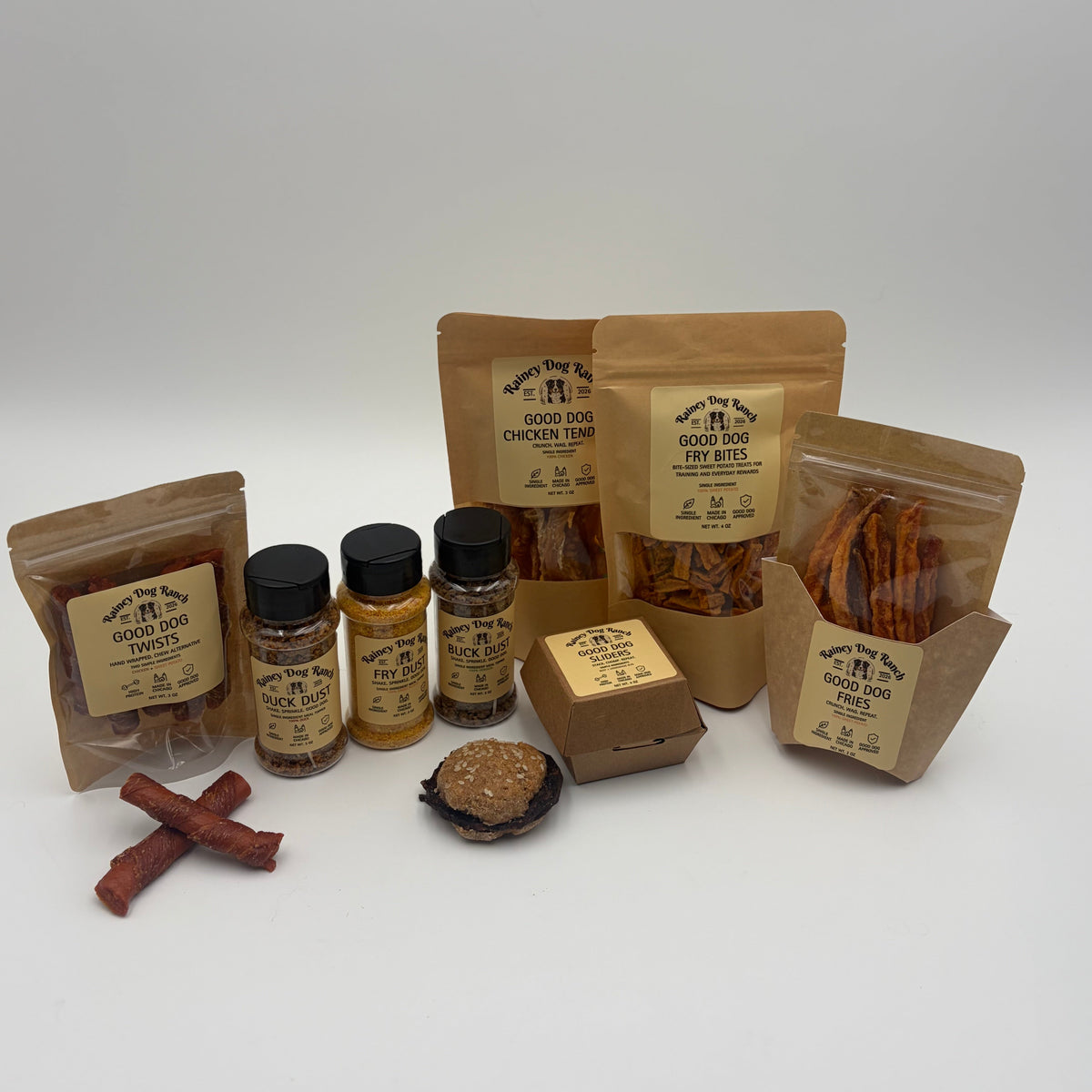 A variety of dog treats and supplements from The Dog Store, including pouches of chicken treat sticks, a jar of joint support supplement, a small box of chow, and a bone-shaped treat, arranged on a white studio background.