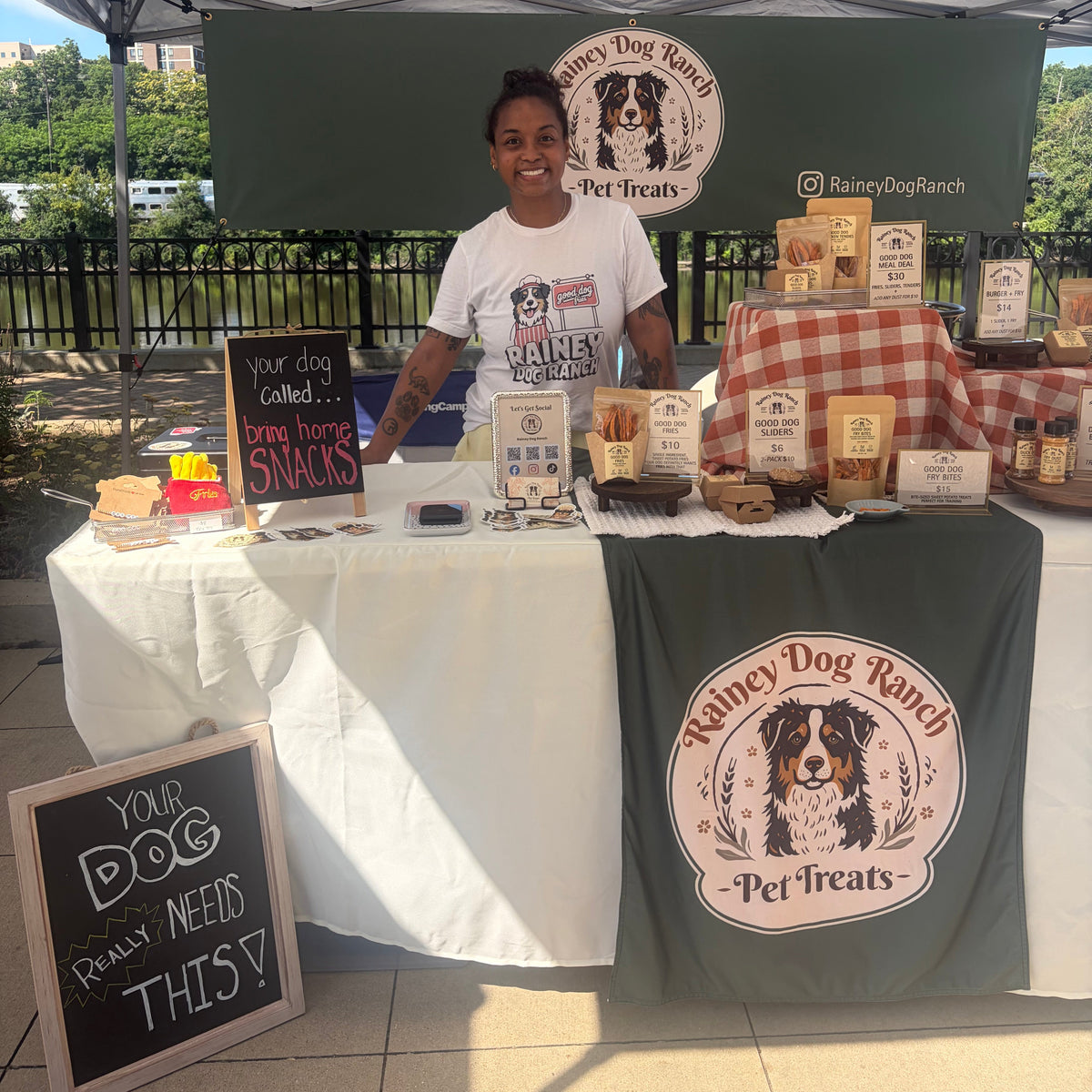 A woman stands behind a market-style booth displaying dog treats from Rainey Dog Ranch. The booth has a white tablecloth, a dark green branded backdrop and drape, and multiple packaged snack products arranged on the table.