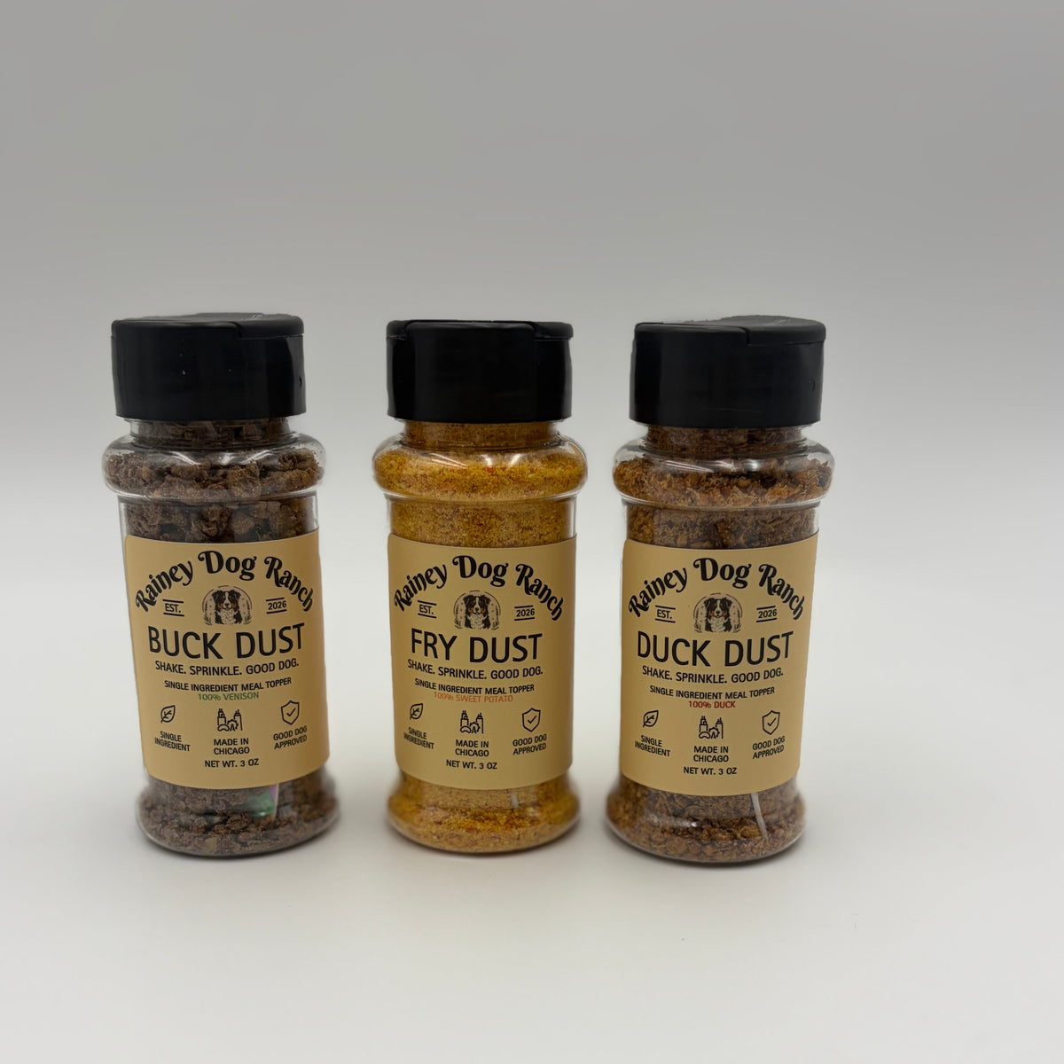 Three clear plastic bottles of Fancy Dog Ranch dog seasoning stands on a white studio background. The bottles contain brown and golden-brown spice dusts labeled Buck Dust, Fry Dust, and Duck Dust, with black caps.