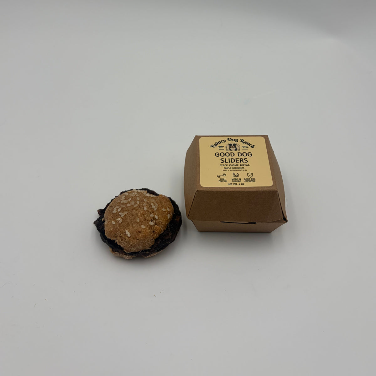 A small brown kraft paper box with a beige label sits on a plain white surface, with a dog hamburger treat beside it
